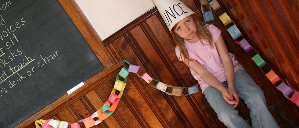 En genrebild av en flicka som sitter i en stol bredvid svarta tavlan, med en dumstrut på huvudet