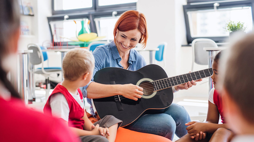 En genrebild av en lärare som sitter på golvet med en grupp barn, läraren spelar gitarr och har ögonkontakt med ett av barnen. Hon ser glad ut