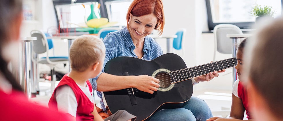 En genrebild av en lärare som sitter på golvet med en grupp barn, läraren spelar gitarr och har ögonkontakt med ett av barnen. Hon ser glad ut