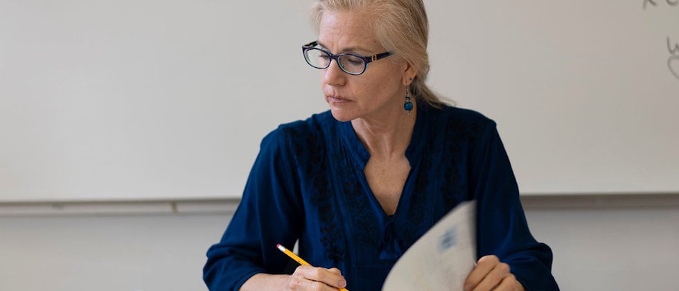 En lärare sitter och arbetar vid sin kateder, hon ser allvarlig ut. I bakgrunden en whiteboard med anteckningar